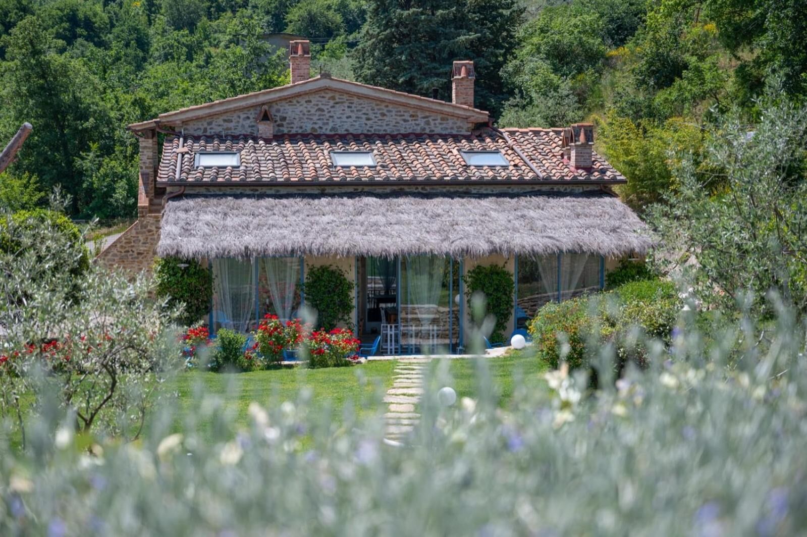 A stone house with a tiled roof and a thatched roof extension, surrounded by lush greenery.
