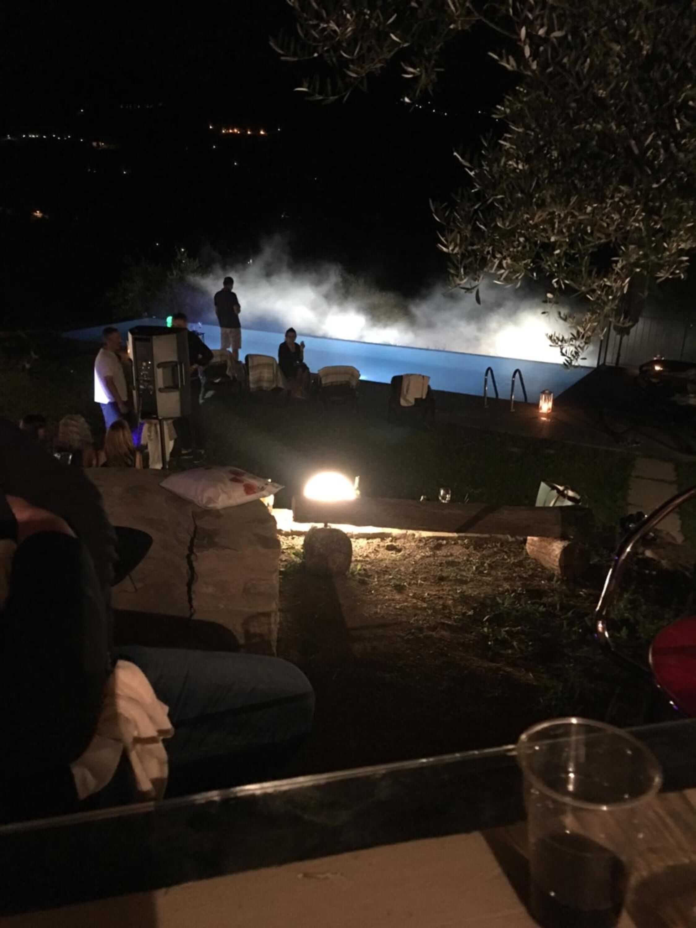 An outdoor seating area with stone tables and chairs, a small stage with a light, and a view of a body of water in the background.
