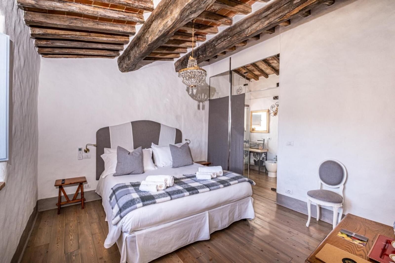 A bedroom with a large bed, a wooden ceiling, a chandelier, a bedside table, a chair, and a mirror.