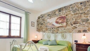 Une chambre avec un mur en pierre, un lit avec une literie verte, une table de chevet avec une lampe et une fenêtre avec des rideaux.
