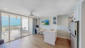 Una cocina moderna con refrigerador, un comedor con mesa y sillas, y un gran ventanal corredero que da a un balcón con vista al océano.
