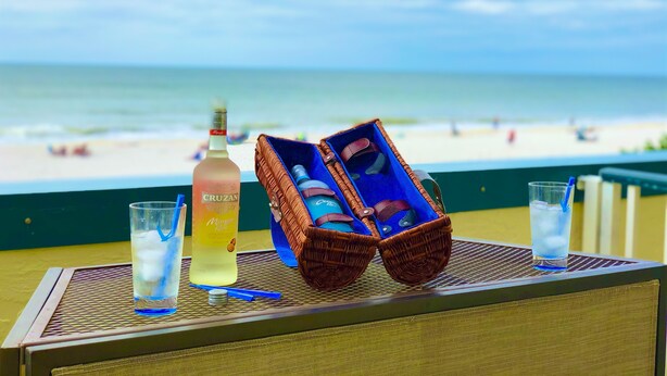 Una mesa con una botella de Cruzan, un vaso de agua y una cesta de mimbre con una botella y un vaso, con vistas a una playa.