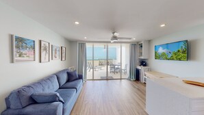 Una sala moderna con un sofá gris, una zona de comedor con mesa y sillas, una cocina con televisor, y un balcón con vista al mar.