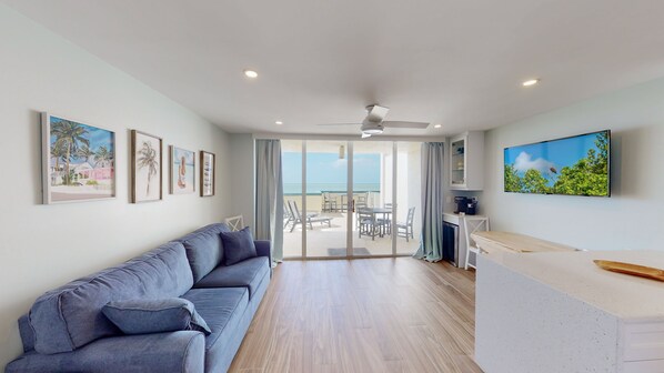 Uma sala de estar moderna com um sofá cinzento, uma área de jantar com mesa e cadeiras, uma cozinha com televisão e uma varanda com vista para o mar.