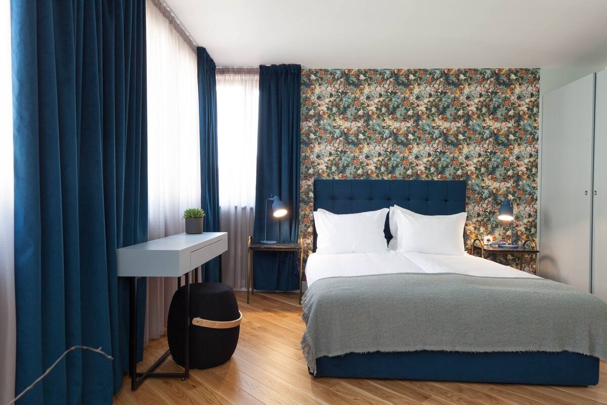 A bedroom with a bed, a desk, a chair, a lamp, and a wall with floral wallpaper.