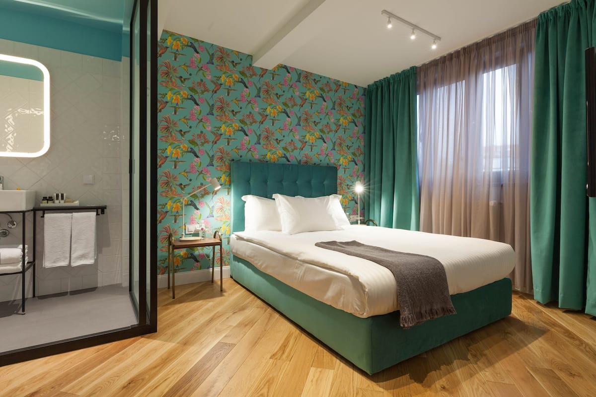 A modern hotel room with a large bed, a bathroom with a mirror, and a colorful floral wallpaper.