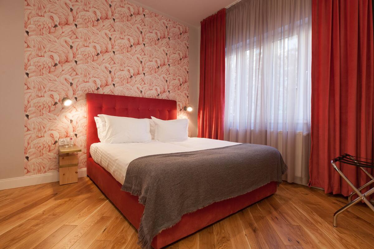 A bedroom with a red bed, white bedding, a red headboard, a wooden nightstand, a red and white striped towel, and a window with red curtains.