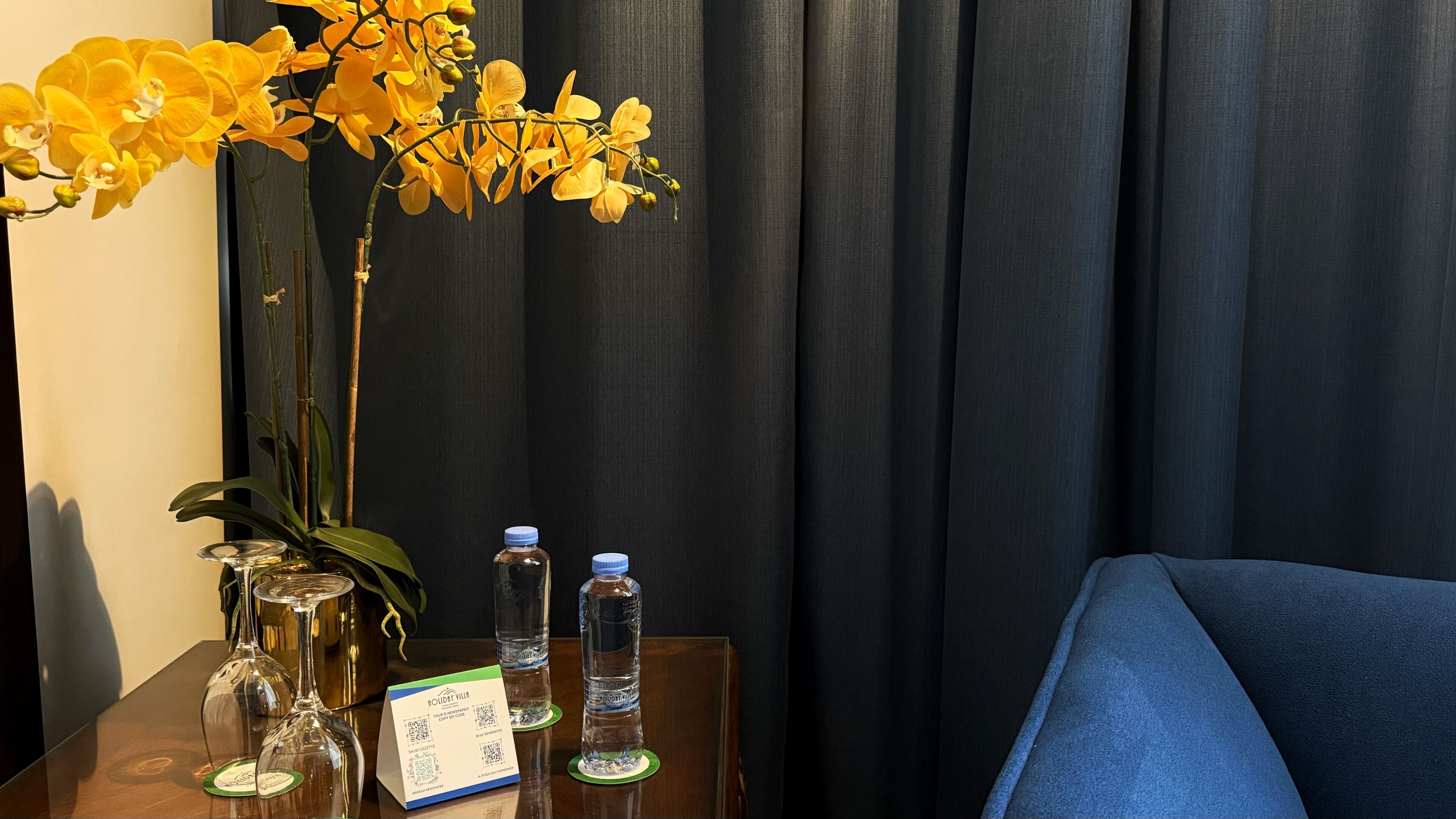 A hotel room with a table featuring a vase of yellow flowers, a wine glass, a water bottle, and a QR code for hotel services.