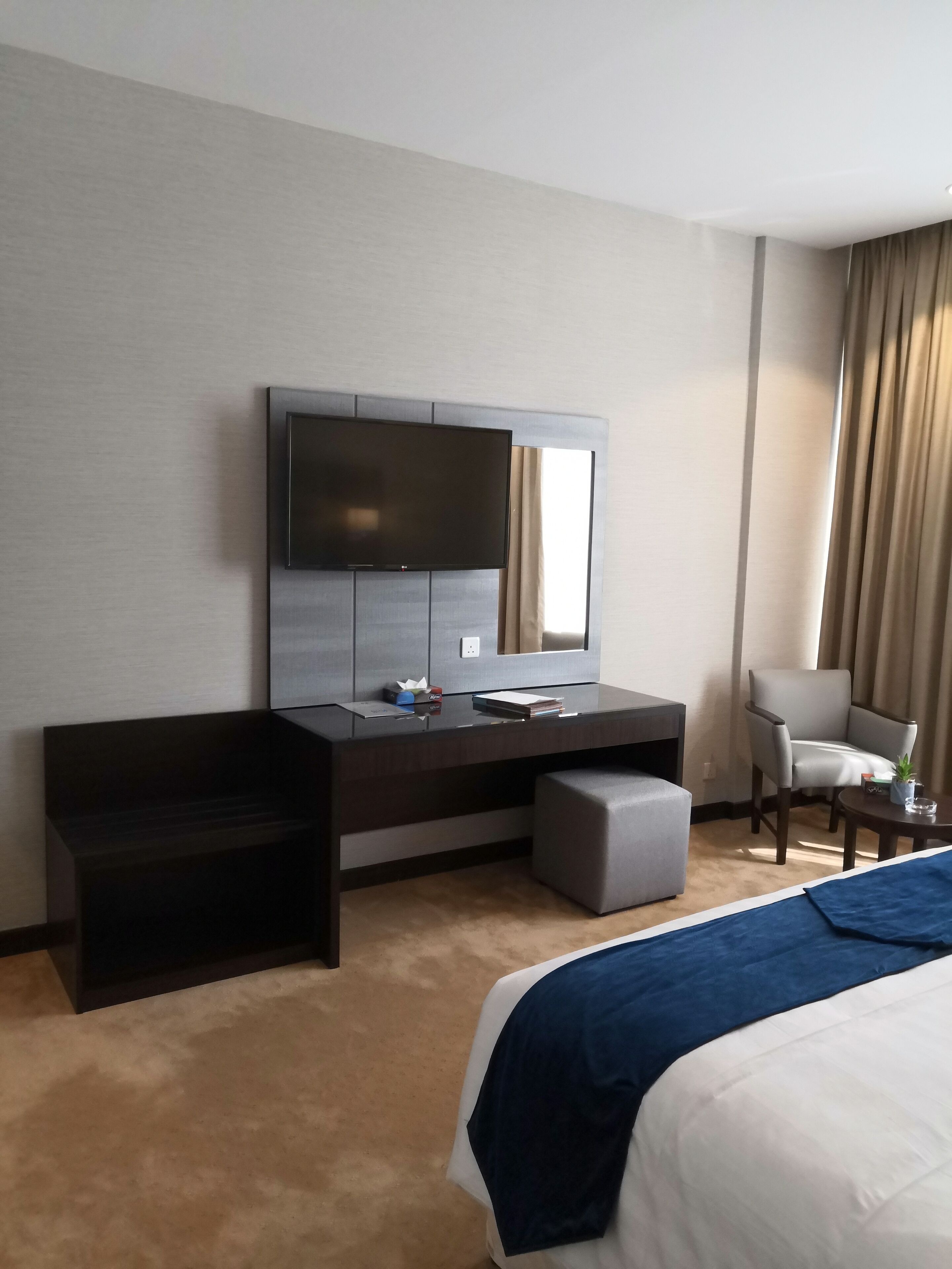 A hotel room with a large bed, a desk with a flat-screen TV, a chair, and a small table.