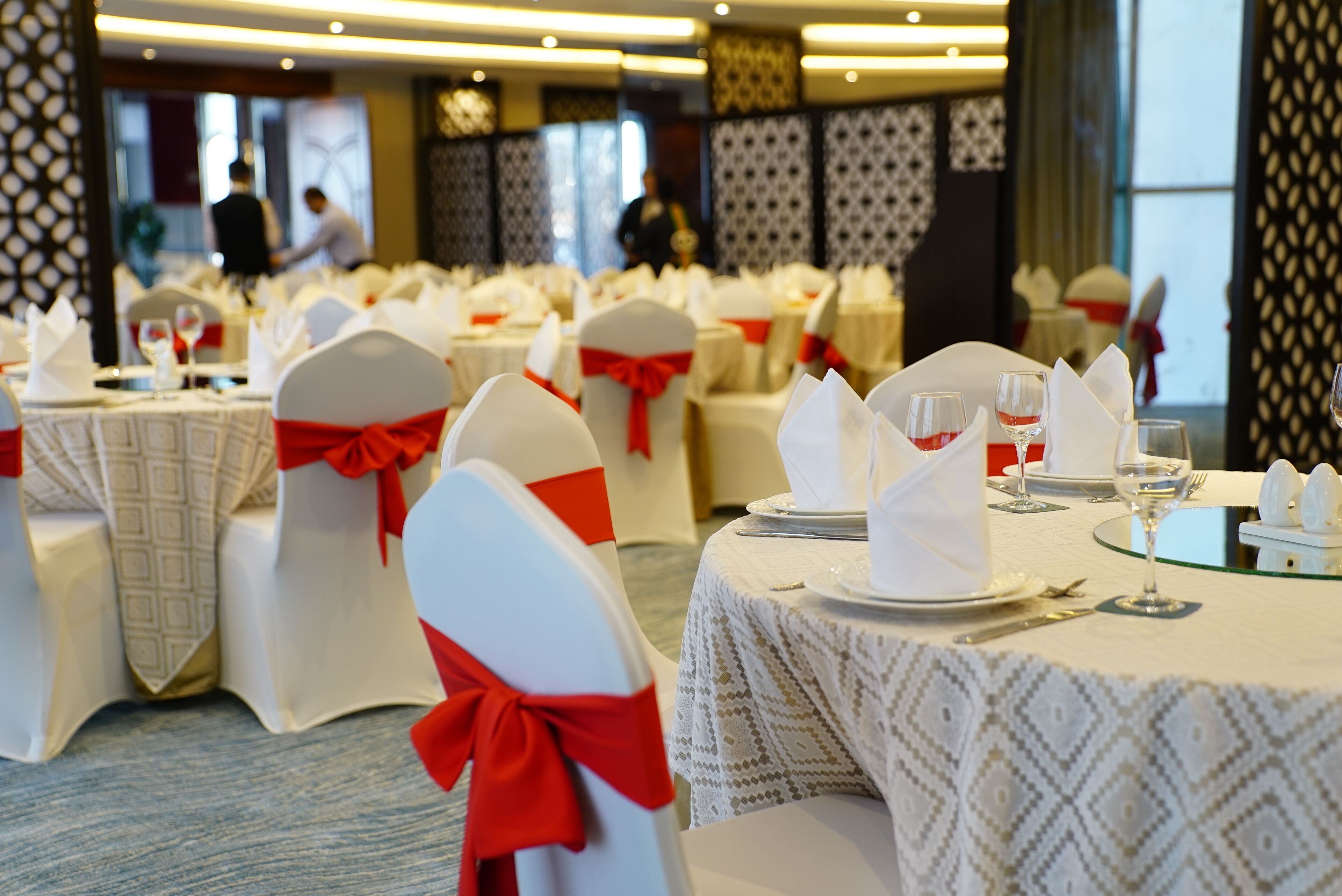 Un salón de banquetes con mesas redondas dispuestas para un evento formal, cada una con manteles blancos y cintas rojas en las sillas.