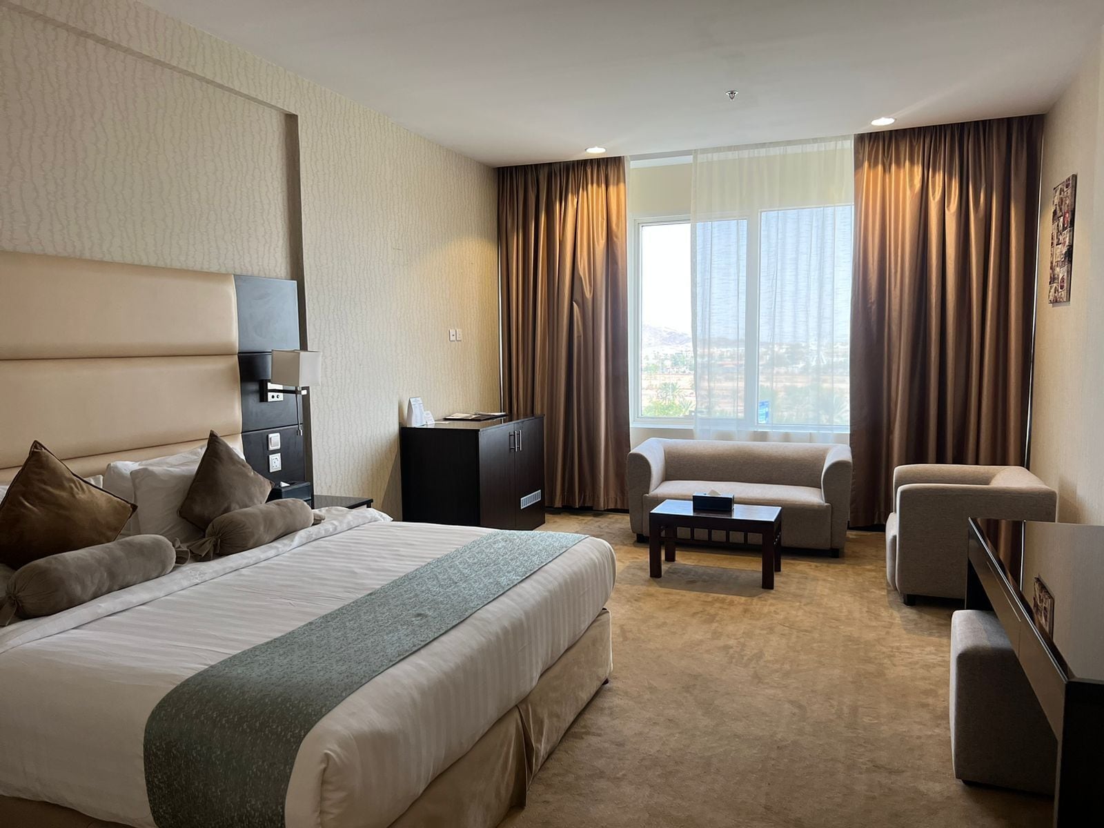 A hotel room with a large bed, a desk, a sofa, and a view of the city through large windows.