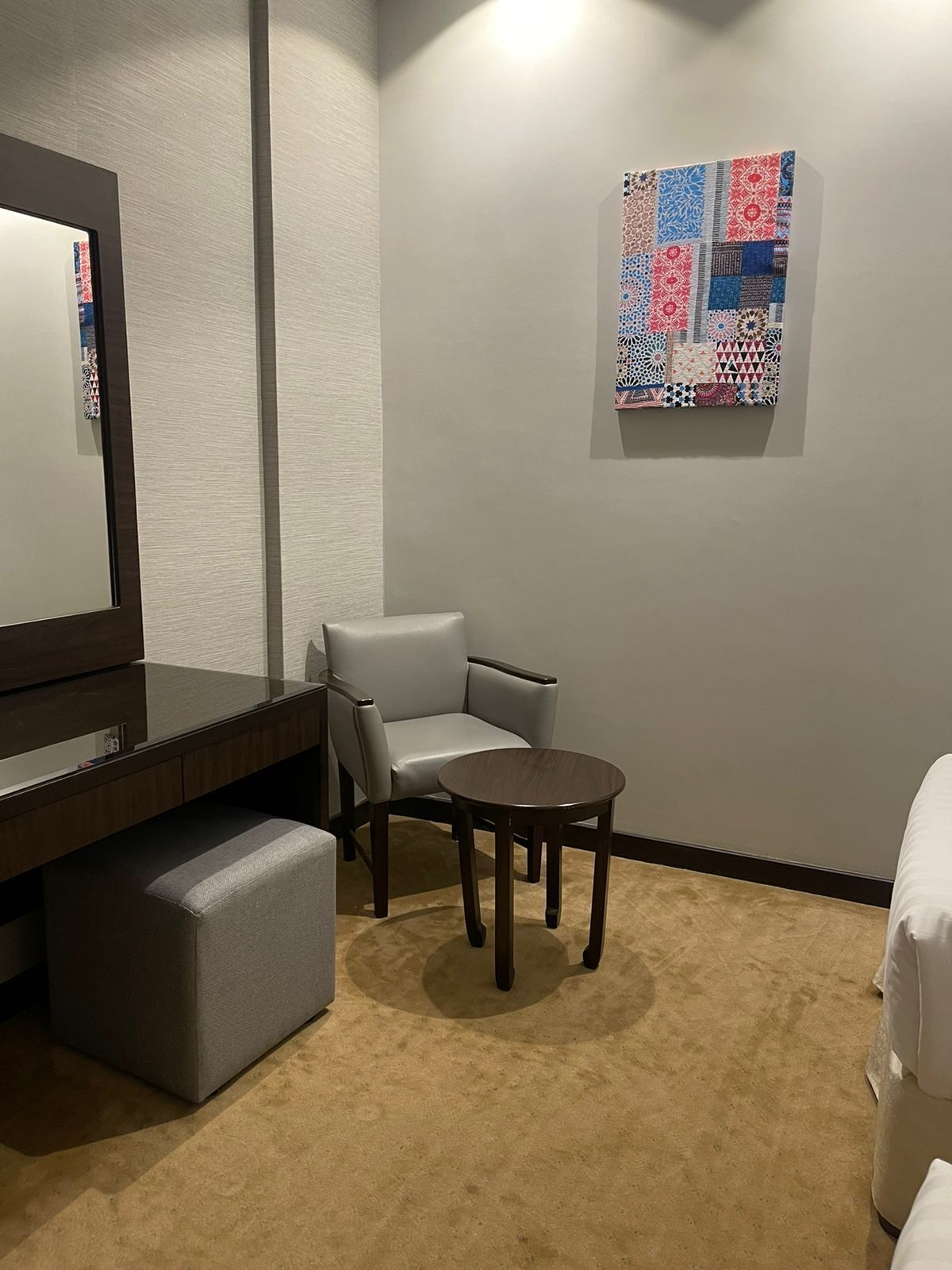 A hotel room with a dark wooden desk, a grey cushioned bench, a small round table, a white armchair, and a colorful abstract art piece on the wall.