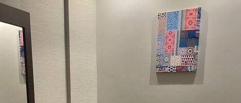 A hotel room with a dark wooden desk, a grey cushioned bench, a small round table, a white armchair, and a colorful abstract art piece on the wall.