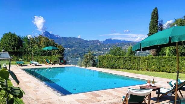 A swimming pool with lounge chairs and umbrellas, surrounded by a landscaped garden and mountains in the background.