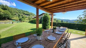 An outdoor wooden dining area with tables set for a meal, surrounded by a lush garden and a mountainous backdrop.