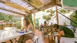 An outdoor patio area with tables and chairs, a thatched roof, and greenery. - Arena Nest Hostel (Santiago del Teide)