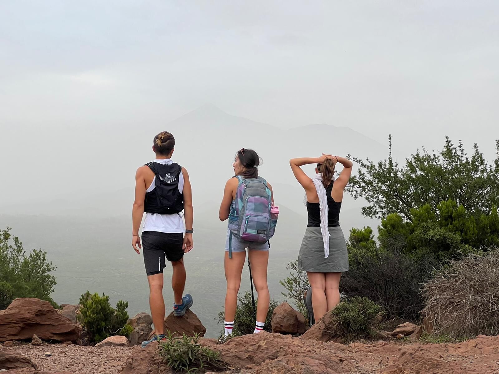 Three people standing on a rocky outcrop overlooking a body of water and mountains.