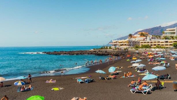 A beach with numerous umbrellas and sun loungers, a rocky breakwater, and a building with multiple balconies overlooking the sea. - Arena Nest Hostel (Santiago del Teide)