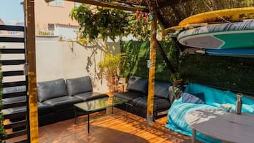 Un patio con un sofá negro, una mesa de vidrio y un sofá azul bajo un techo de paja.