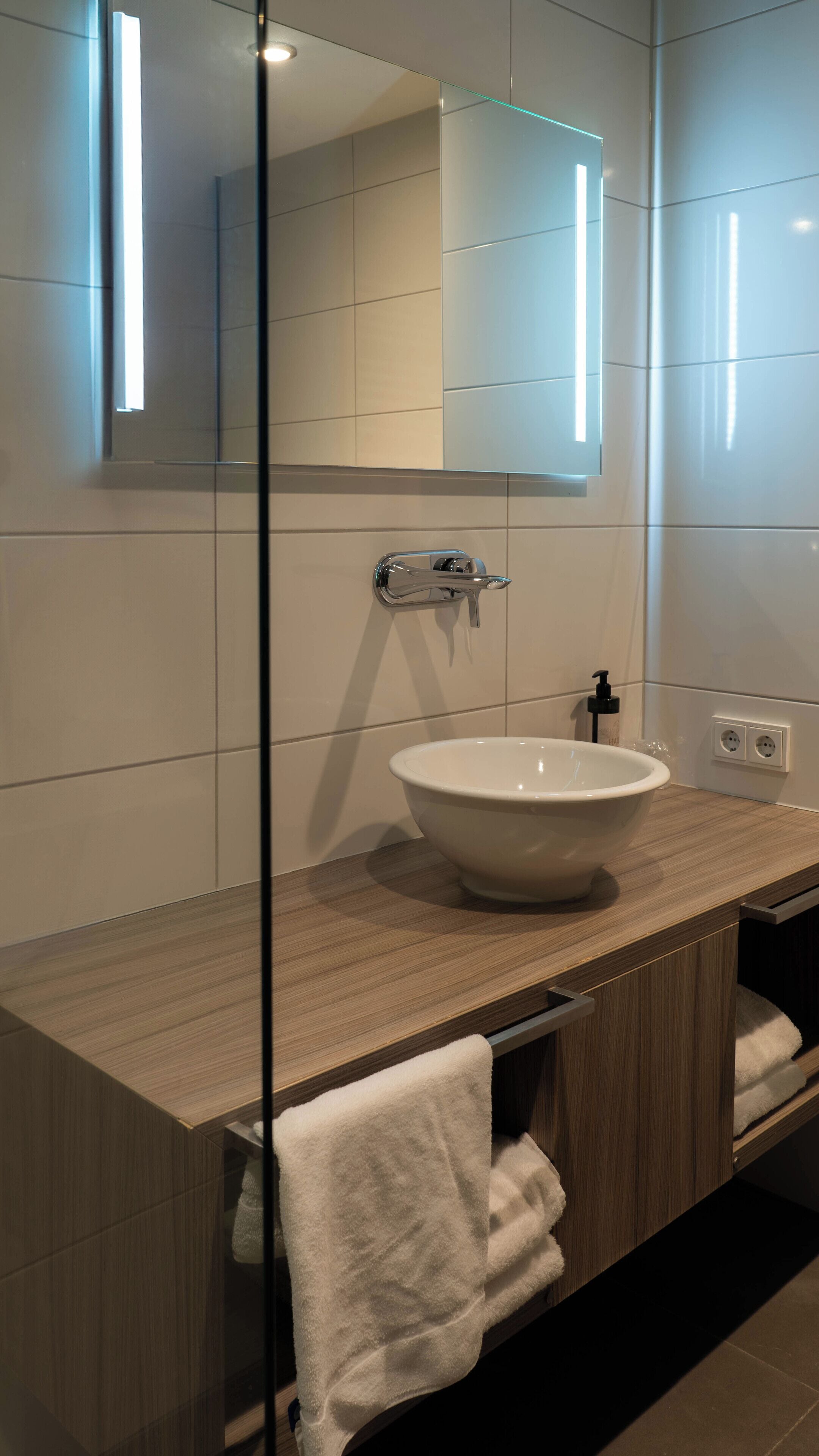 A modern bathroom with a large mirror, a white sink, and a wooden vanity.