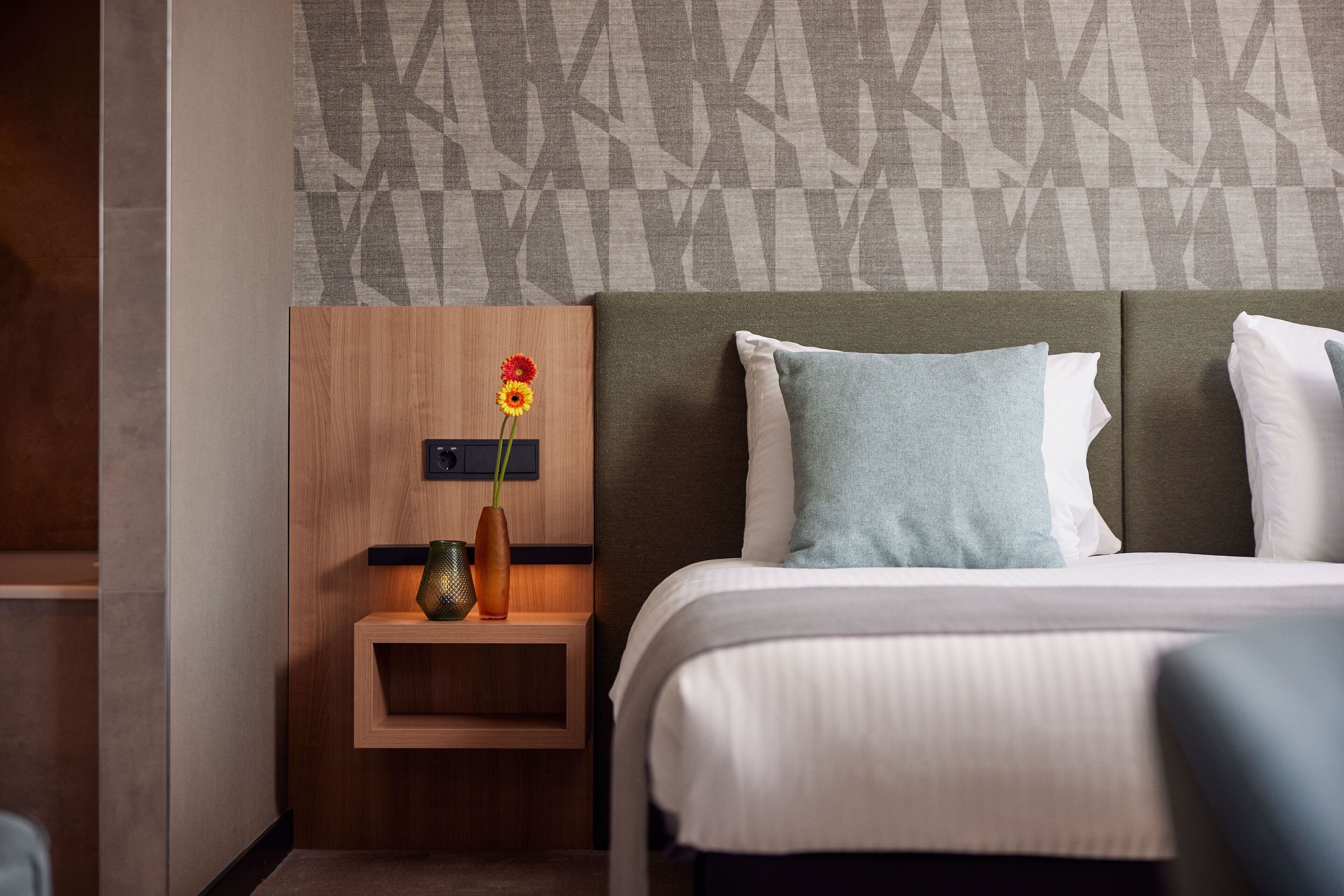 A neatly made bed with a green headboard and white pillows, a small wooden nightstand with a vase of flowers, and a patterned wall in the background.
