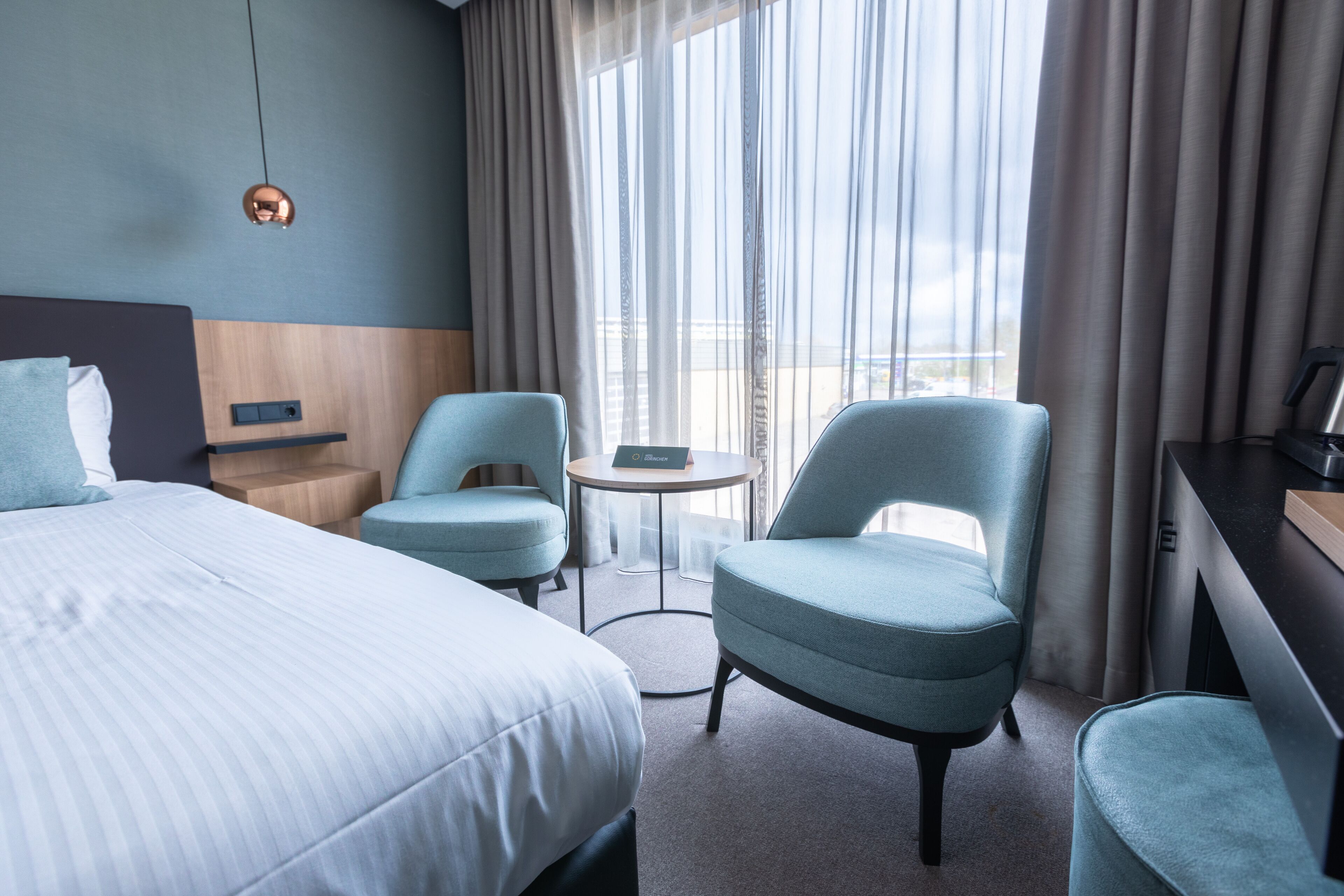 A hotel room with a large bed, two armchairs, a small round table, and a view of the city through sheer curtains.