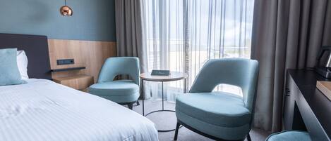 A hotel room with a large bed, two armchairs, a small round table, and a view of the city through sheer curtains.