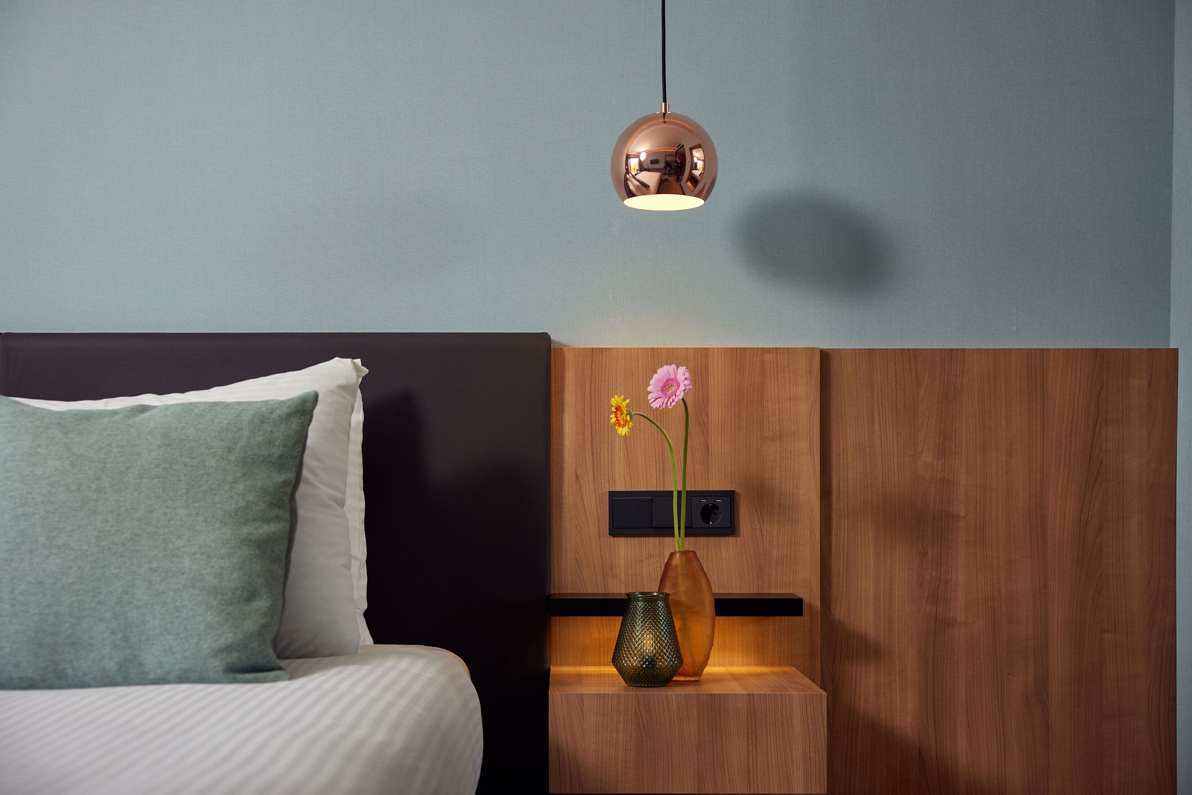 Een hotelkamer met een bed, een nachtkastje met een bloemenvaas en een hanglamp.