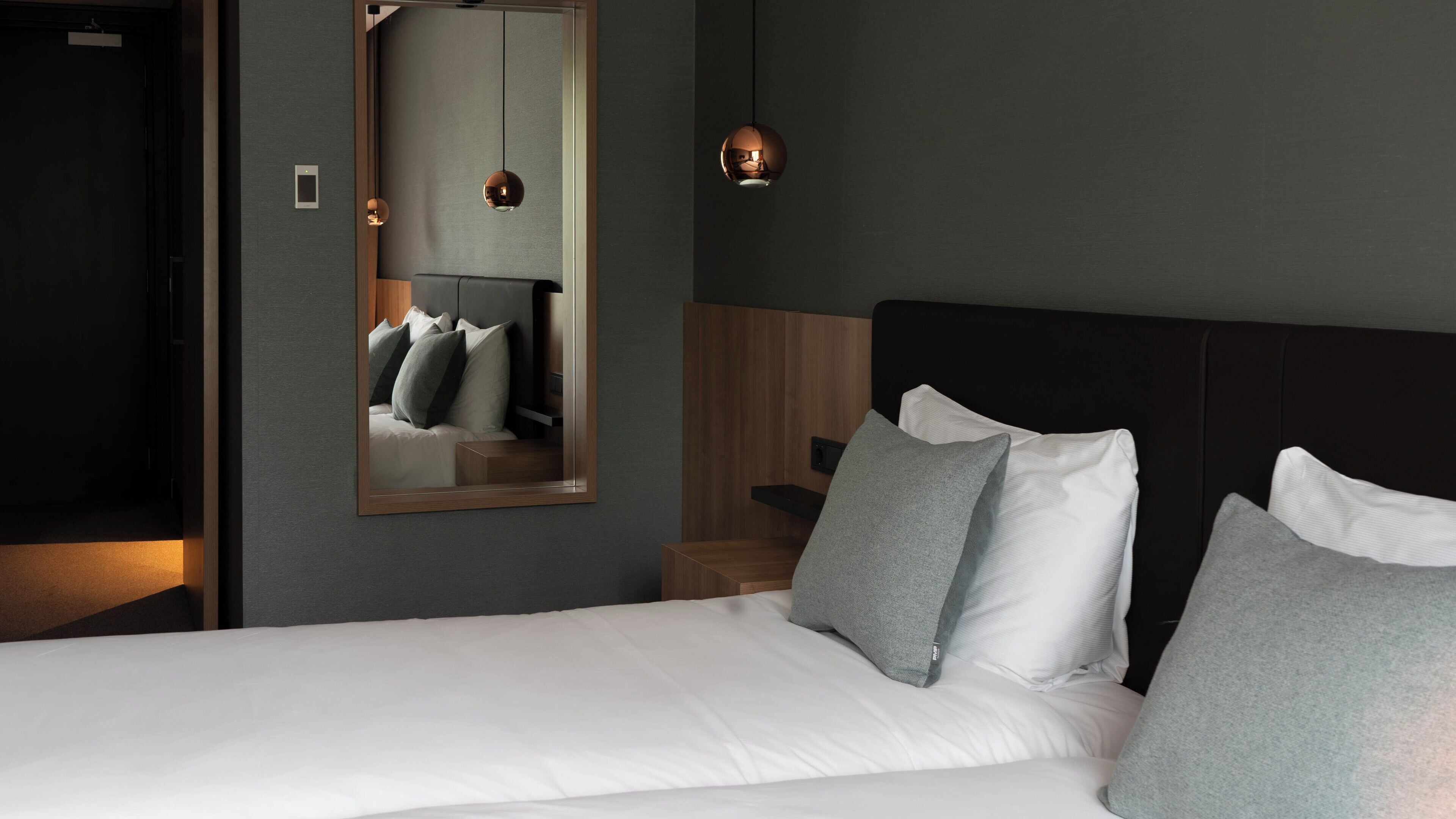 Een keurig opgemaakt bed met witte lakens en grijze kussens, een houten hoofdbord, een nachtkastje met een lamp en een spiegel aan de muur.
