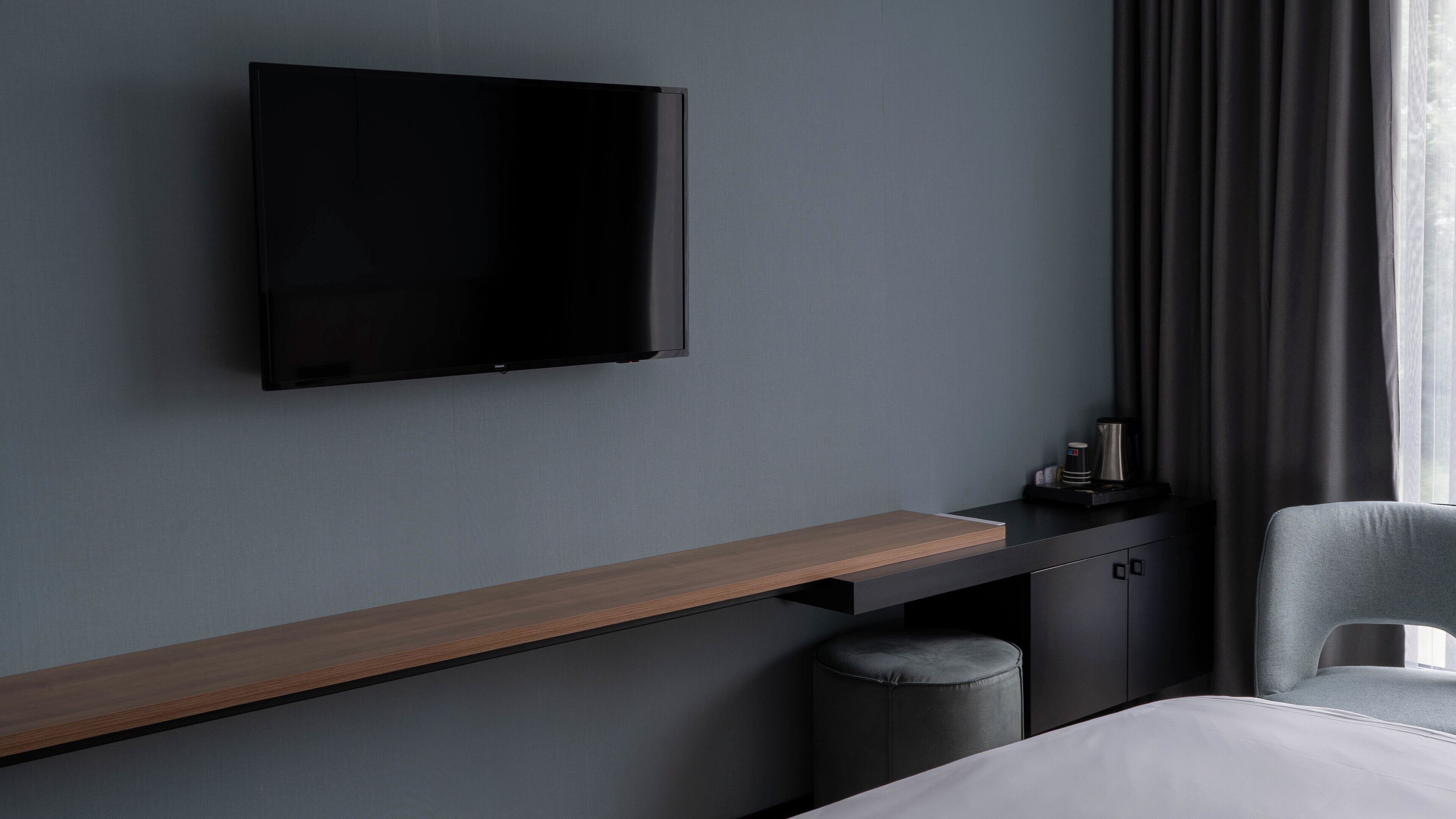 Una habitació d'hotel moderna amb televisió plana penjada a la paret, una taula consola de fusta, una cadira tapissada de color gris i un petit reposapius.