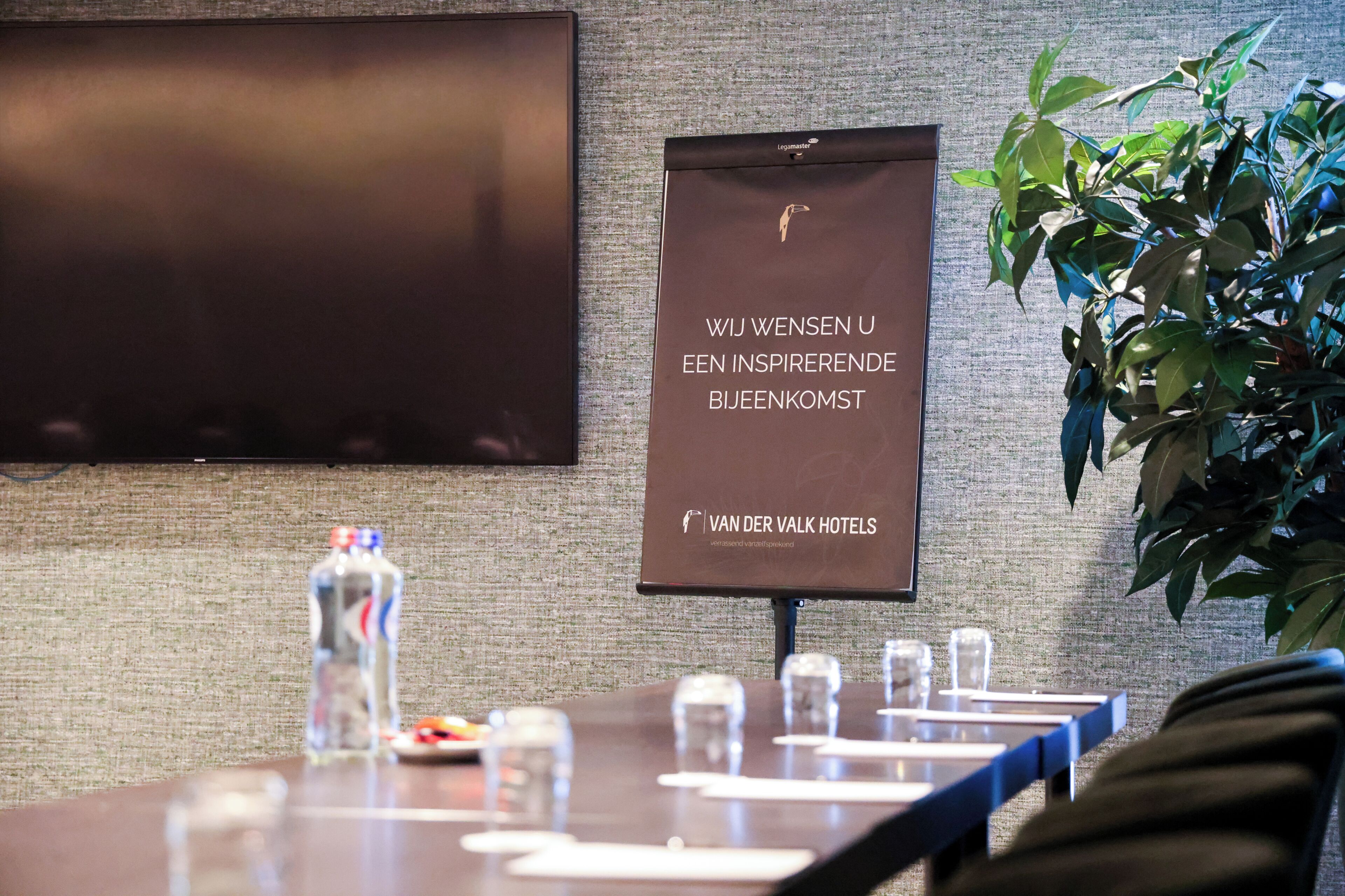 Una mesa de conferencias con botellas de agua y vasos, un cartel con texto y una planta.