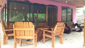 Een overdekt terras met houten stoelen en een bank, grote ramen en een roze muur.