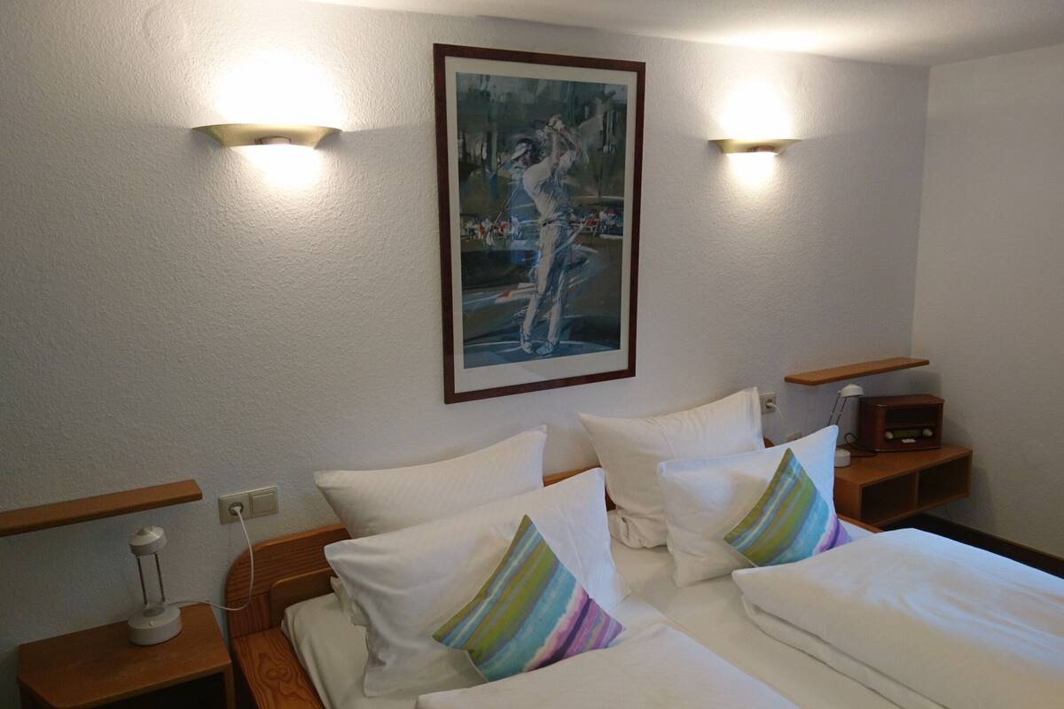 A hotel room with a bed, two bedside tables, a nightstand, a painting on the wall, and two wall-mounted lights.