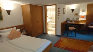 A hotel room with a bed, a desk, a chair, a small table, and a bathroom visible through an open door.