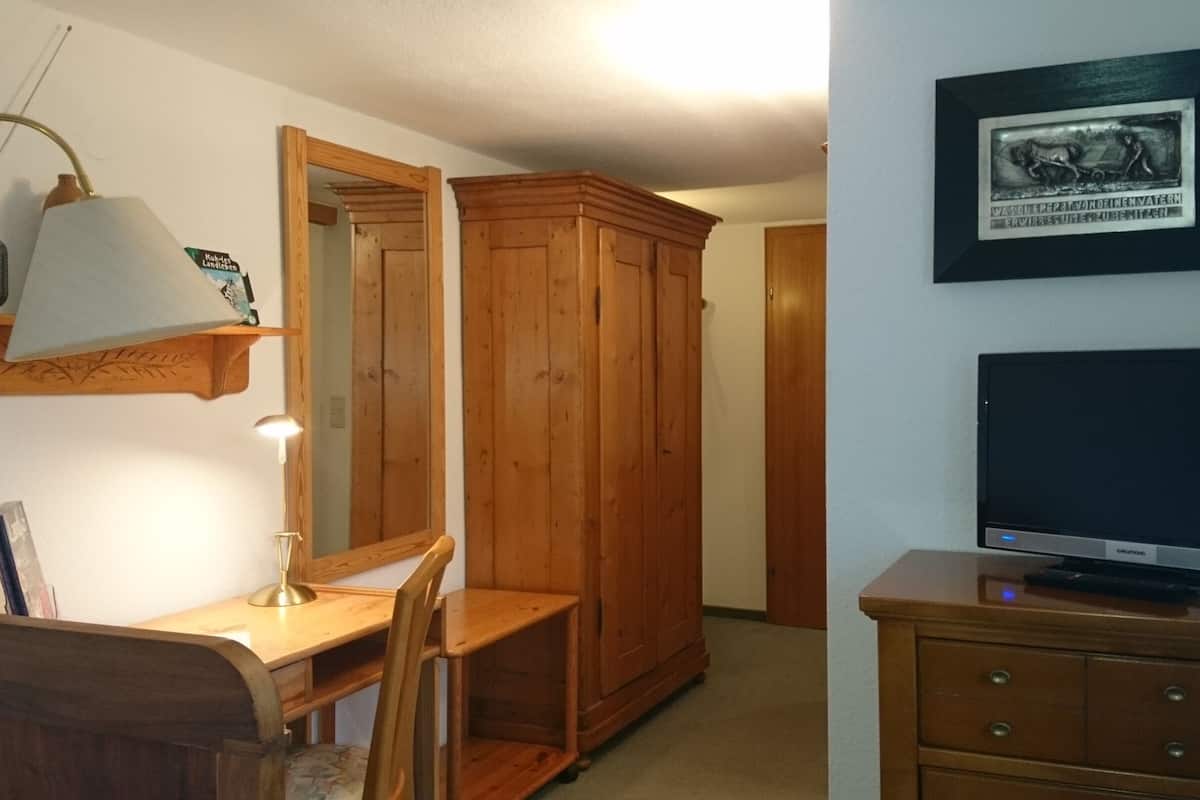 A room with a wooden desk, chair, and lamp; a wardrobe; a television on a stand; and a framed picture on the wall.