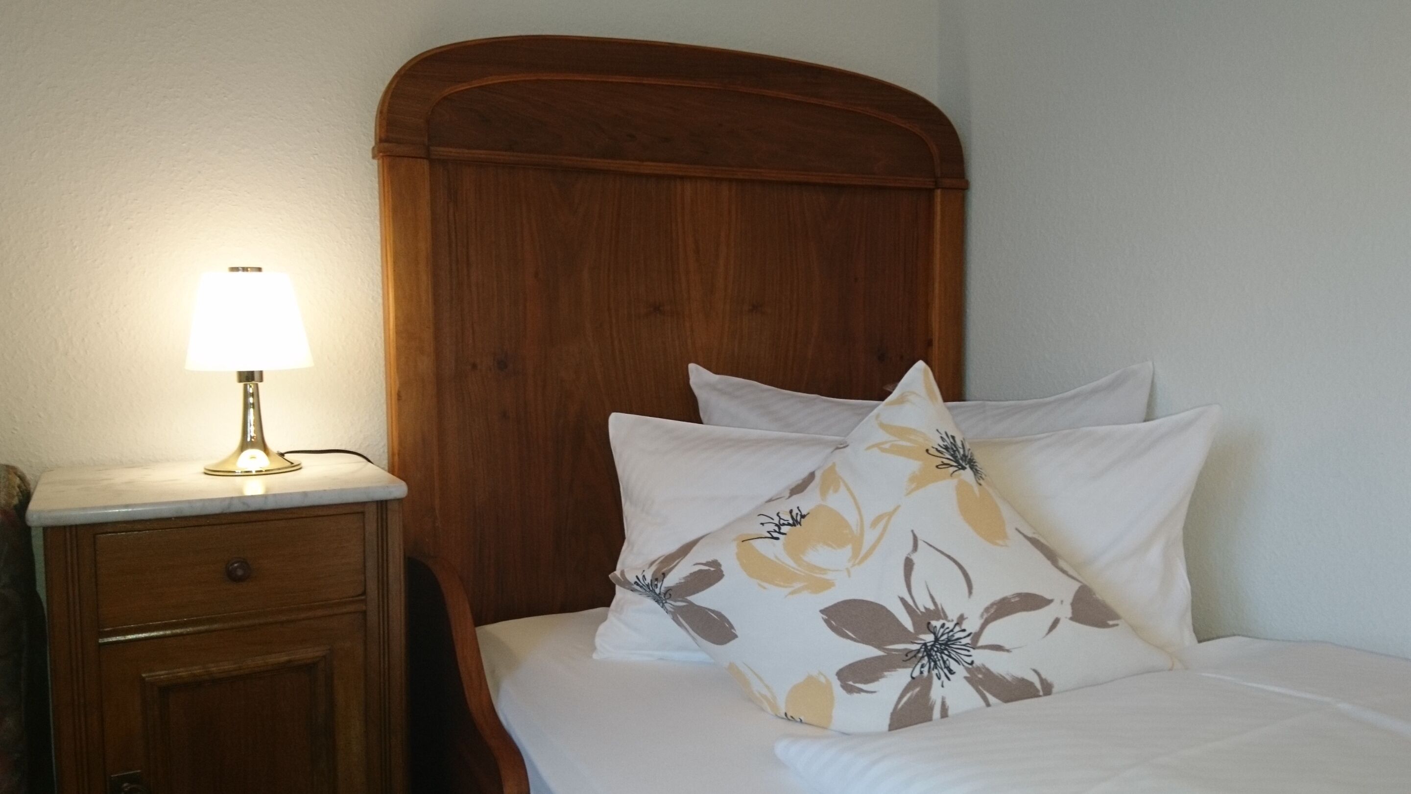 Una cama bien hecha con almohadas estampadas, un cabecero de madera y una lámpara de noche sobre una mesita de mármol.