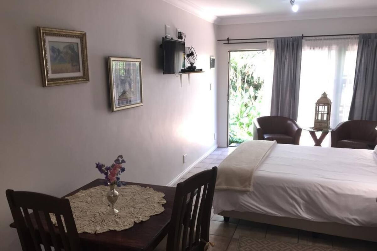 Een slaapkamer met een bed, een tafel met stoelen, een bijzettafeltje met een lantaarn en een raam met gordijnen.