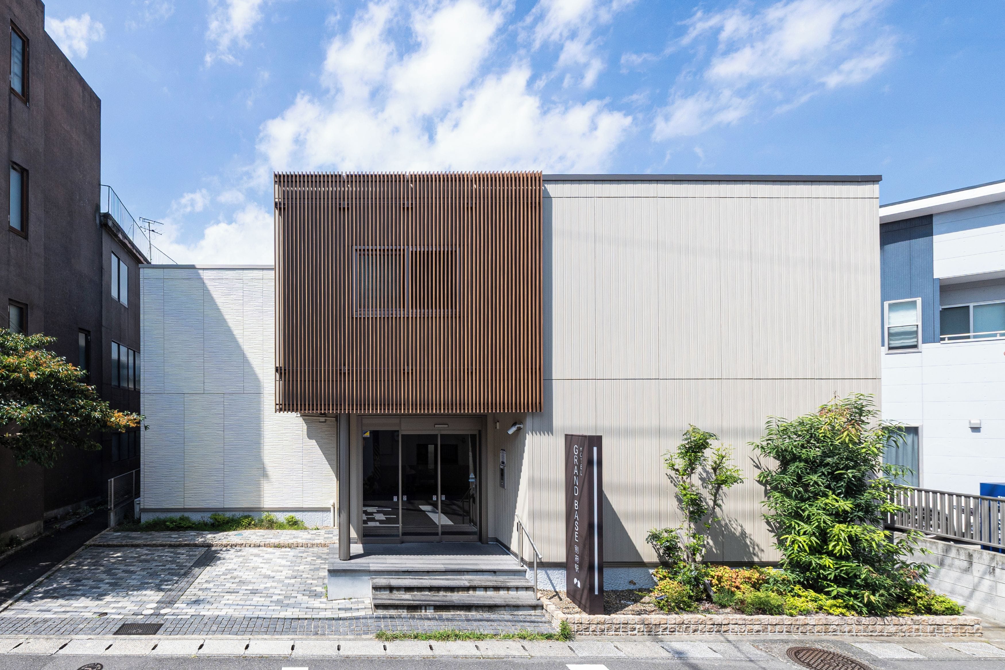 A modern building with a vertical wooden panel facade, a glass entrance, and a sign indicating 'Hotel Grand Base.'
