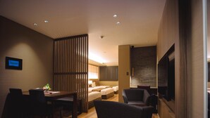 Ein modernes Hotelzimmer mit Holzboden, einer Essecke mit Tisch und Stühlen, einem Bett mit Kopfteil, einem an der Wand befestigten Fernseher und einer Trennwand mit vertikalen Lamellen.