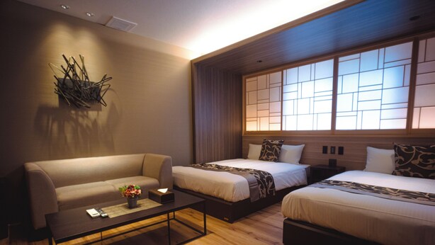 Ein modernes Hotelzimmer mit einer Couch, zwei Betten, einem Couchtisch und einem großen Fenster mit traditionellen Jalousien.