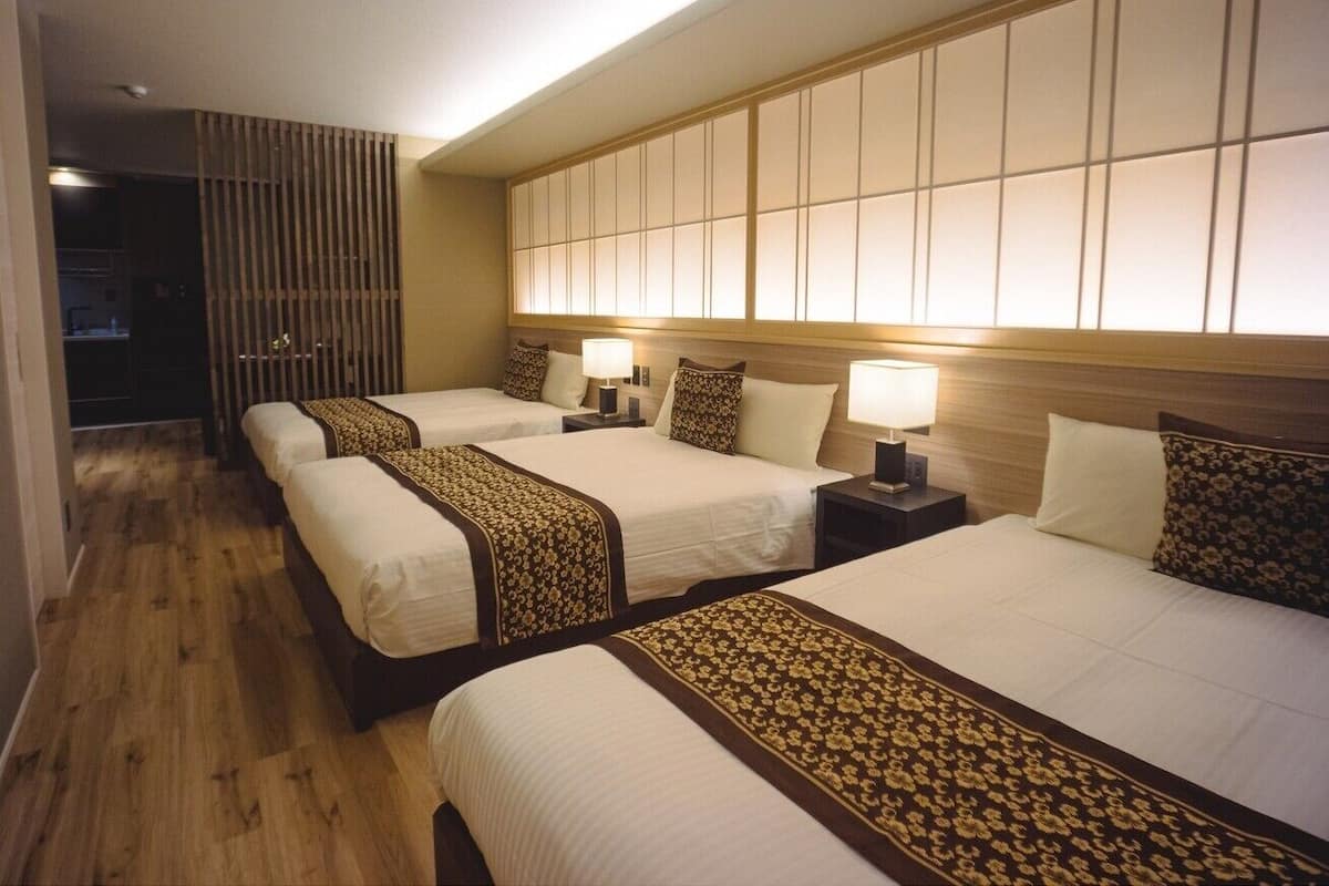 Habitación de hotel con tres camas, piso de madera y un ventanal grande con persianas.