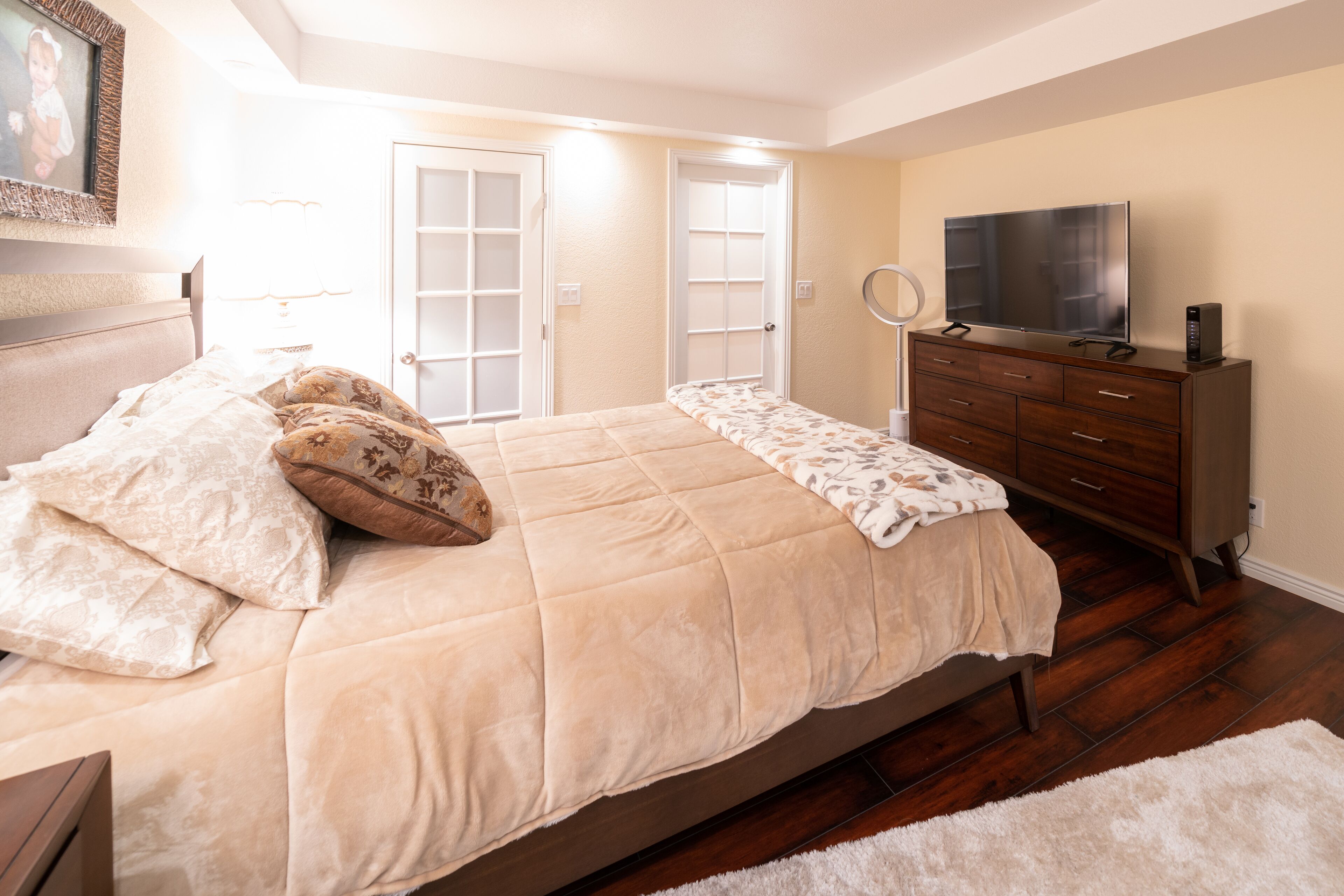 A bedroom with a large bed, a dresser, a TV, and a door leading to another room.