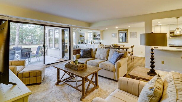 A spacious living room with a large sliding glass door leading to an outdoor area, a cozy seating arrangement, and a kitchen area visible through an open door. - 1421 South Beach Villa is a beachy 4 bedroom, 3 bath townhome (Hilton Head Island)