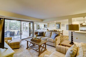 A spacious living room with a large sliding glass door leading to an outdoor area, a cozy seating arrangement, and a kitchen area visible through an open door.