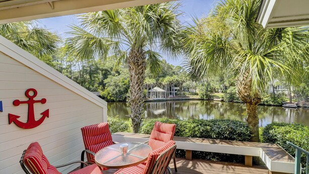 A deck with a glass table and striped chairs, overlooking a pond with palm trees and a gazebo. - 122 Abbington is a beautiful 3-bedroom, 3 bath villa in Palmetto Dunes (Hilton Head Island)