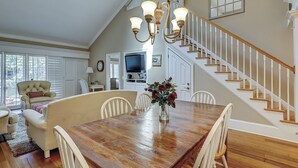 A spacious dining area with a wooden table, white chairs, and a chandelier. A staircase with white railings leads to an upper level. - 122 Abbington is a beautiful 3-bedroom, 3 bath villa in Palmetto Dunes (Hilton Head Island)