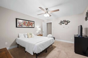 Quarto com cama, ventilador de teto, TV na parede e porta para outro cômodo.