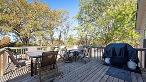 Una terraza de madera con una mesa, sillas, una parrilla y árboles al fondo.