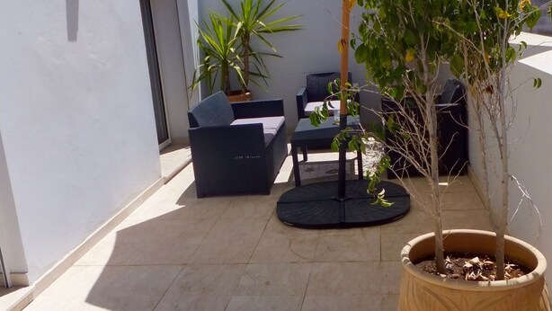Uma área de pátio com um grande guarda-sol, mobiliário de exterior e plantas em vasos.