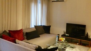 Uma sala de estar com sofá, mesa de centro, televisão e janela com cortinas.